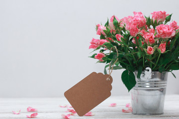 Bunch of roses in tin bucket, petals and paper tag for message on white wooden table. Women day...