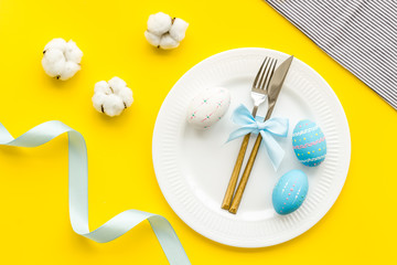 Easter table decoration. Plate, cutlery, painted eggs and dry white flowers, tablecloth on yellow background top view