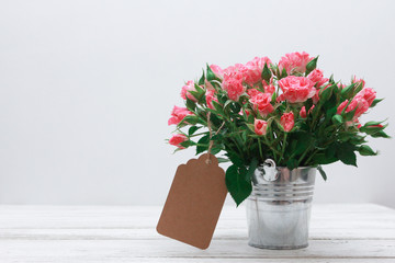 Bunch of roses in tin bucket and paper tag for message on white wooden table. Women day concept. Spring, 8 march, wish, copy space, craft