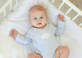 Charming smiling blue-eyed 9 month old baby in a gray bodysuit is lying in a child's bed. Top view