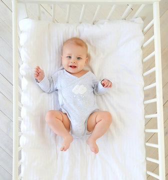 Charming smiling blue-eyed 9 month old baby in a gray bodysuit is lying in a child's bed. Top view