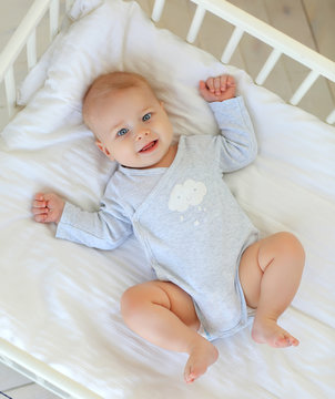 Charming smiling blue-eyed 9 month old baby in a gray bodysuit is lying in a child's bed. Top view