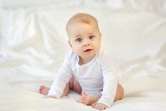 Charming blue-eyed 9 month old baby in white clothes sitting on the bed