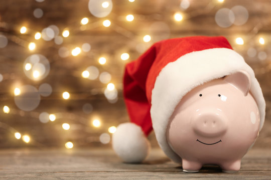 Piggy bank with Santa hat on table against blurred Christmas lights. Space for text