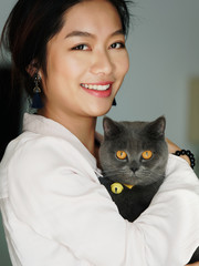 Portrait of beautiful Chinese young girl hug her cute British Shorthair Cat who has amazing orange eyes at home and smile at camera in sunny afternoon, happy pets owner lifestyle.