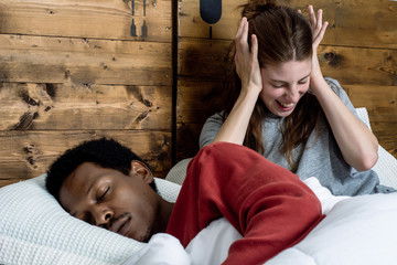 Woman blocking ears with hands while man snoring on bed