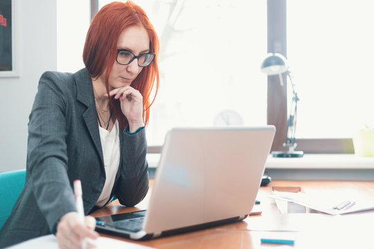 A Ginger Woman In Glasses Sitting At Office And Looking At The Laptop