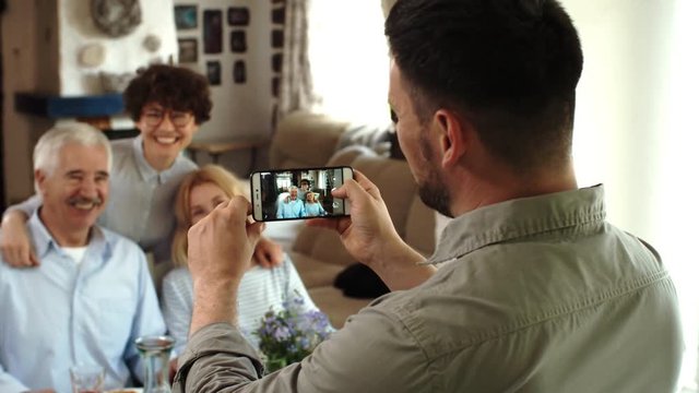 Medium shot of man standing with smartphone in his hands, making photo of young woman and her parents