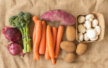 Top down view of loose organic vegetables from farmers market on hessian background, including onions, carrots, broccoli, sweet potato, potatoes and mushrooms