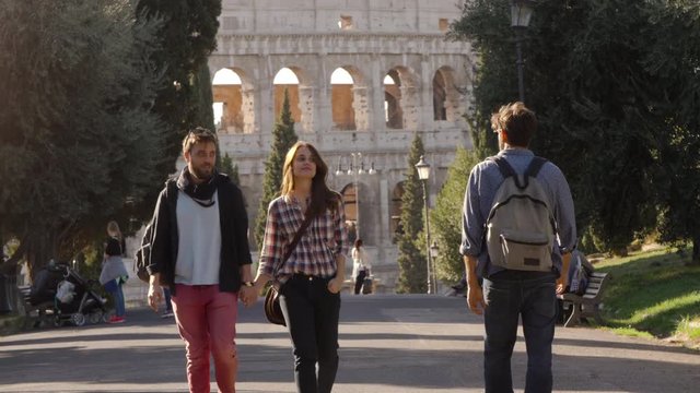 Young happy couple walk in park road and attractive young man glance to girlfriend gelousy with trees colosseum in background in rome at sunset lovely beautiful girl with long hair holding hands slow