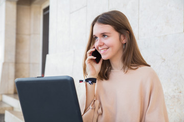 Smiling teenage girl using laptop and phone at building wall