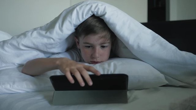 A boy closeup is watching cartoons on a digital tablet at night under a blanket. Concept of electronic addiction