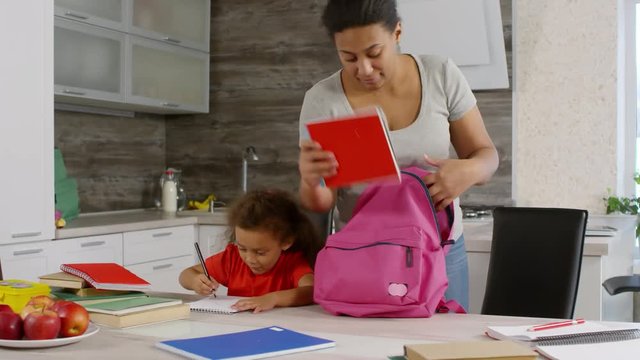 African girl sitting at table and drawing when her mother coming in with school bag and starting packing it, and then helping girl putting it on. Little girl going away and her mother waving to her
