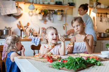 Children are eating italian homemade pizza. Cute kids are having fun while enjoying delicious food in cozy home kitchen. Three girls at family dinner table. Lifestyle, authentic moment.