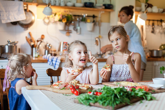 Children Are Eating Italian Homemade Pizza. Cute Kids Are Having Fun While Enjoying Delicious Food In Cozy Home Kitchen. Three Girls At Family Dinner Table. Lifestyle, Authentic Moment.