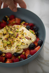 Frau isst Beeren Obstsalat mit Erdbeeren, Himbeeren, Blaubeeren, Pistazien, Zitronen Zesten, Leinöl Kurkuma Quark Creme in blauer Schale als Frühstück auf Marmor Hintergrund