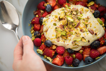 Frau isst Beeren Obstsalat mit Erdbeeren, Himbeeren, Blaubeeren, Pistazien, Zitronen Zesten, Leinöl Kurkuma Quark Creme in blauer Schale als Frühstück auf Marmor Hintergrund