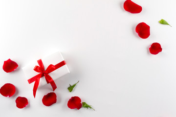White gift box with red ribbon red velvet rose petals on white background. Flat lay