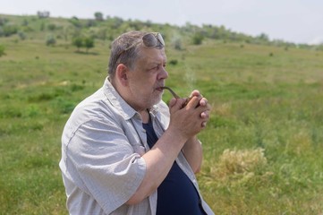 Naklejka na ściany i meble Nice portrait of happy Ukrainian countryman smoking tobacco pipe while sitting outdoor against hilly spring pasture