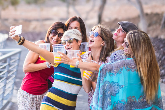 Group of happy and cheerful caucasian young women toasting and clinking with plastic glasses and taking a selfie picture in friendship and having fun all together in outdoor leisure activity