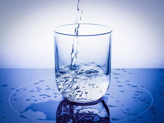 A jet of water pours into a glass with bubbles and drops on a blue background.