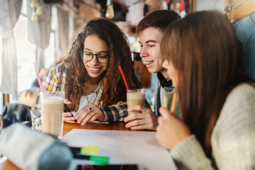 Three smiling multicultural classmates sitting at coffee shop, drinking coffee and looking at smart...