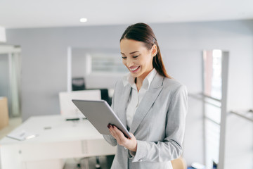 Smiling Caucasian female CEO in formal wear and with long brown hair using tablet while standing at office. Small opportunities are often the beginning of great achievements.