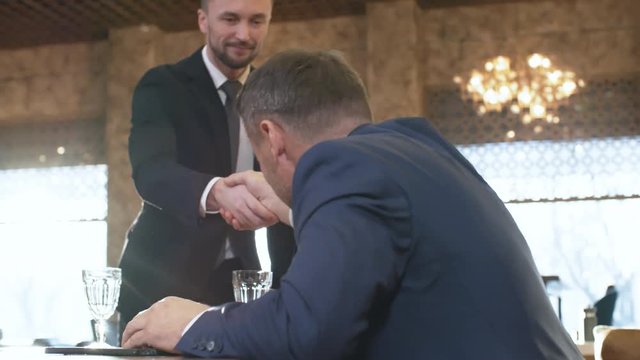 Panning of middle-aged businessman sitting at table, then his male partner approaching. They greeting each other by shaking hands and starting talking