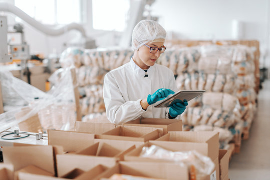 Young Caucasian employee in sterile uniform using tablet for logistic. Food factory interior.