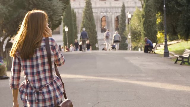 Beautiful girl walks in park road talking on phone and attractive young man boyfriend behind her taps shoulder run funny prank trees colosseum in background in rome at sunset lovely beautiful woman