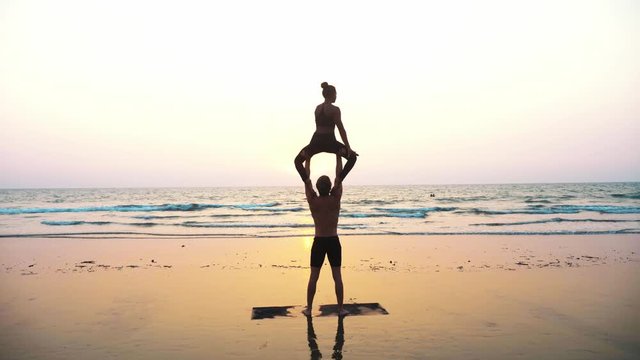 Silhouette of fit sporty couple practicing acrobatic yoga with partner together on the beach. Steadicam shot of male standing and holding his female partner over his head.