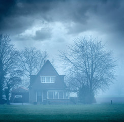 House with winter trees in mist under cloudy sky.
