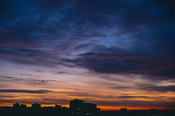 Cityscape with vivid warm dawn. Amazing dramatic blue violet cloudy sky above dark silhouettes of city buildings. Orange sunlight. Atmospheric background of sunrise in overcast weather. Copy space.
