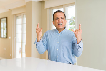 Middle age man sitting at home shouting with crazy expression doing rock symbol with hands up. Music star. Heavy concept.