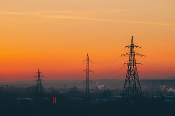 Power lines in city on dawn. Silhouettes of urban buildings among smog on sunrise. Cables of high voltage on warm orange yellow sky. Power industry at sunset. City power supply. Mist urban background.