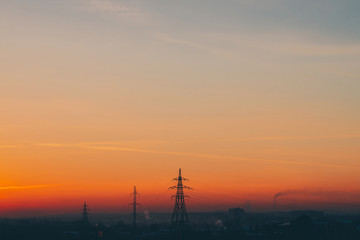 Fototapeta premium Power lines in city on dawn. Silhouettes of urban buildings among smog on sunrise. Cables of high voltage on warm orange yellow sky. Power industry at sunset. City power supply. Mist urban background.