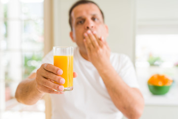 Middle age man drinking a glass of orange juice at home cover mouth with hand shocked with shame for mistake, expression of fear, scared in silence, secret concept