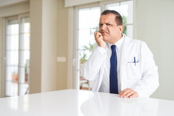 Fototapeta premium Middle age doctor man wearing medical coat at the clinic looking stressed and nervous with hands on mouth biting nails. Anxiety problem.