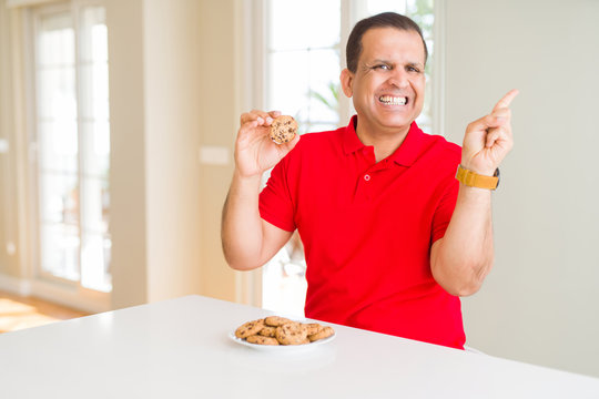 Middle age man eating chocolate chips cookies at home very happy pointing with hand and finger to the side