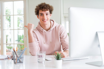 Young student man using computer pointing and showing with thumb up to the side with happy face smiling