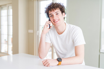 Young man talking on the phone at home with a happy face standing and smiling with a confident...