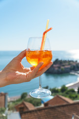 A man is holding an orange cocktail in a glass with ice and tubules on the background of European houses with red tiles, the Adriatic Sea and the island of Sveti Stefan.