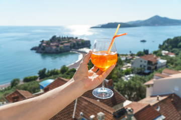 A man is holding an orange cocktail in a glass with ice and tubules on the background of European houses with red tiles, the Adriatic Sea and the island of Sveti Stefan.
