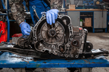 A close-up of a young man repairman in a working uniform of cars is repairing an automatic gearbox...