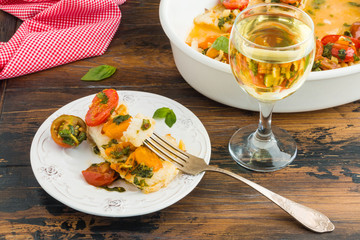 Roasted cod fillet with cherry tomato and basil sauce in white dish and on plate on wooden rustic table, glass of wine