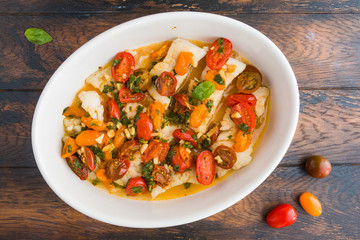 Fried cod fillet with cherry tomato and basil sauce in white dish on wooden rustic table, top view