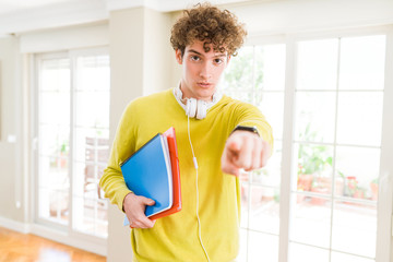 Young student man wearing headphones and holding notebooks pointing with finger to the camera and to you, hand sign, positive and confident gesture from the front