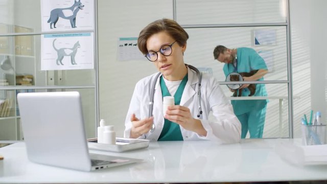 Lockdown of female veterinarian sitting at computer in medical office, explaining client how to use pills by video call, while her male assistant examining cat in funnel cone on background