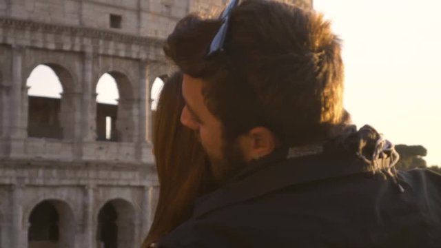 Romantic young couple tourists doing famous TITANIC scene in front of colosseum in rome at sunset boyfriend holding girlfriend spread arms happy beautiful woman long hair slow motion sun flair