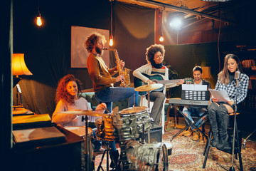 Jazz band preparing for the gig. In foreground woman singing while the rest of the band playing bass guitar, clavier and acoustic guitar. Home music studio interior.